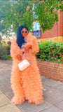 Woman in a peach fur coat standing on a sidewalk with greenery and a building in the background.