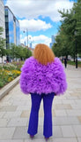 Person wearing a purple exaggerated couture coat and blue pants walking on a city street.