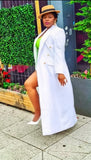 Woman in a white coat and hat standing on a sidewalk with greenery in the background