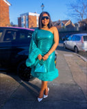 Woman in a teal dress with a feathered shrug standing on a street.