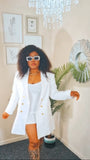Woman in a white blazer and dress standing in a room with decor elements.